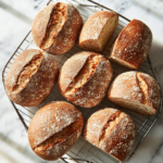 Freshly baked multi-grain bread loaves being removed from pans and placed on a cooling rack, with a knife slicing into one loaf to reveal the soft, airy crumb inside.