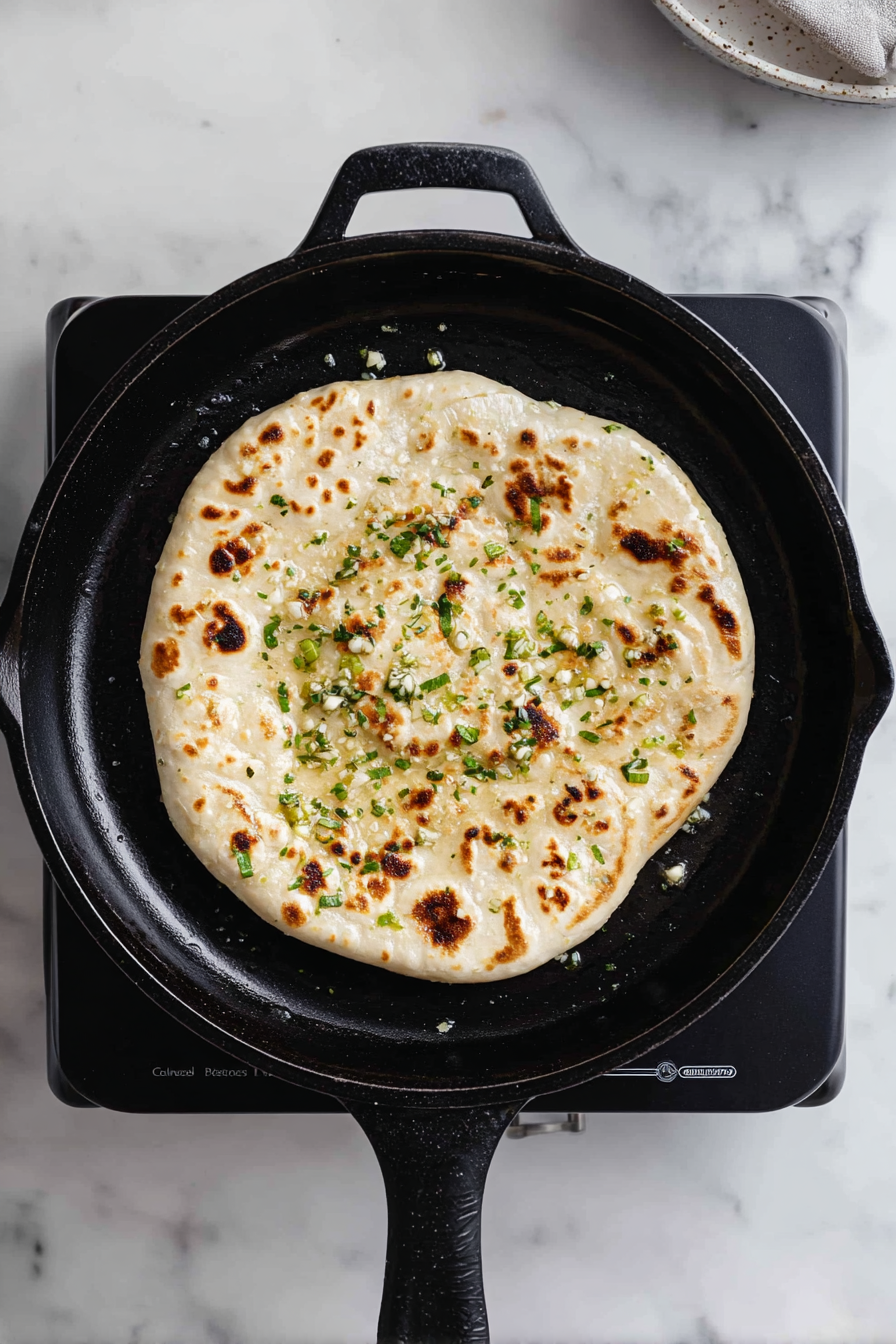 Heat cast-iron skillet over moderately high heat, press minced garlic into the dough, brush with melted butter, and cook each naan for about 1 minute until puffy.