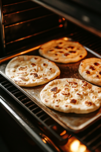 A top-down view of flatbreads baking in an oven, turning golden brown with a light crisp forming on the edges. The oven door is slightly open, with light from the oven highlighting the texture of the flatbreads.