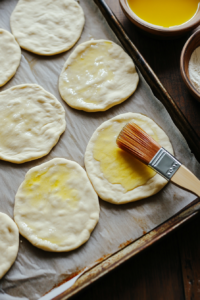 A top-down view of rolled-out flatbreads on a parchment-lined baking sheet, being brushed with olive oil. The light is warm and natural, highlighting the smooth texture of the dough and the sheen from the olive oil.