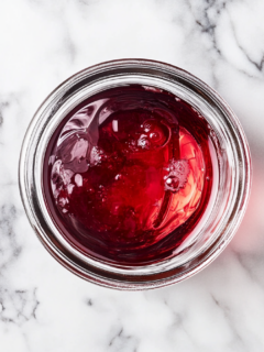 Clear cherry-infused liquid being poured into a 1-quart jar on a marble countertop, ready for storage with the sediment left behind.