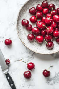 Fresh cherries being pricked with a knife tip on a marble countertop. Thawing frozen cherries with their juices reserved in a small bowl.