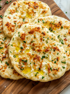 A top-down view of freshly baked cottage cheese flatbreads on a wooden board or platter on a marble countertop. The flatbreads are golden brown with a light, crispy texture, drizzled with olive oil or sprinkled with fresh herbs, and the scene is clean and inviting.