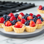 This image shows the completed mini fruit tarts with golden brown tart shells filled with smooth pastry cream and garnished with fresh strawberries and blueberries, making for a visually appealing and delicious dessert.