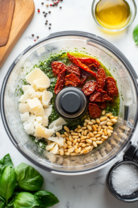 The image shows a top-down view of a food processor with sun-dried tomatoes, fresh basil, Parmesan cheese, pine nuts, and garlic inside. Salt, pepper, and olive oil are nearby, and the pesto is being blended. The scene is well-lit to highlight the rich colors and textures of the pesto.