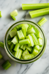 The image shows a top-down view of celery being washed and sliced into ½-inch pieces on a cutting board. The sliced celery is neatly packed into a wide-mouthed 16-ounce jar, ready for pickling. The light highlights the fresh, crisp texture of the celery.