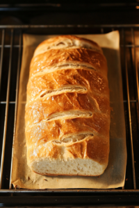 The image shows a loaf of ciabatta warming in the oven on a baking sheet, with a golden-brown crust forming. After warming, the ciabatta is sliced horizontally through the center. The light captures the texture of the bread as it heats.