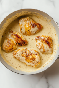 The image depicts cooked chicken pieces being added to the thickened yogurt sauce in a saucepan. The scene captures the melding of flavors with warm, natural light.