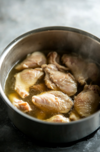 The image shows a large pot on a stovetop with chicken pieces covered in salted water, gently simmering. The scene focuses on the simmering process with soft, natural light.