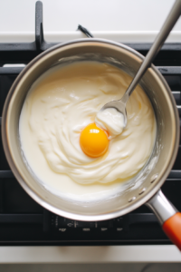 The image shows the saucepan with yogurt sauce on the stovetop, being thinned with cooking liquid from the chicken. The rich, creamy consistency of the sauce is highlighted by soft, natural light.