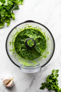 Fresh parsley, cilantro, garlic, red wine vinegar, and olive oil being blended in a food processor to create a vibrant chimichurri sauce for the rice.