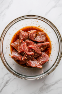 Flank steak slices being marinated in a flavorful mixture of soy sauce, grated ginger, garlic, sugar, and mirin inside a clear bowl, ready for refrigeration to absorb all the rich flavors.