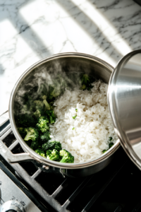 A covered saucepan simmering white rice and broccoli in broth, showing the cooking process for the Creamy Broccoli Cheddar Rice Recipe.