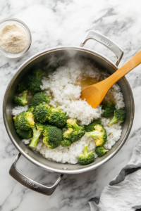 A wooden spoon stirring white rice and broccoli florets into boiling broth in a medium saucepan, as part of the Creamy Broccoli Cheddar Rice Recipe.