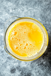 Slowly pouring warm milk into the egg yolk and sugar mixture to temper the eggs, ensuring a smooth consistency for the Natilla pudding.