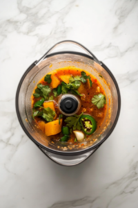 This image shows the ingredients for the marinade, including orange juice, onion, jalapeños, garlic, cilantro, and spices, being blended in a high-speed blender to create a smooth marinade.