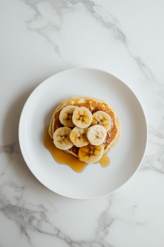 This image shows warm banana pancakes topped with fresh banana slices and drizzled with syrup, served on a simple plate set against a marble cooktop background.