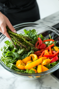 Roasted asparagus and chopped baby bell peppers being added on top of the fresh greens and cabbage in the salad bowl