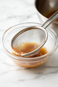 Warm apple juice mixture being strained through a fine-mesh strainer into a bowl, removing spices like cinnamon, cloves, and star anise to create a smooth liquid for the Non-Alcoholic Spiced Apple Fizz.