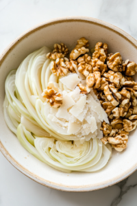 Thinly sliced fennel in a serving bowl topped with shaved Parmesan and half the toasted walnuts forming the base of the salad