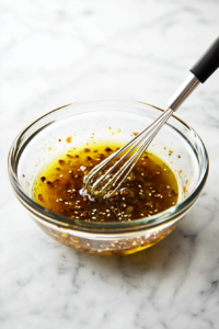 This image shows making the dressing for a chicken salad. Garlic, ginger, vegetable oil, Dijon mustard, toasted sesame oil, rice vinegar, lemon juice, soy sauce, salt, sugar, and pepper are whisked together in a small bowl.