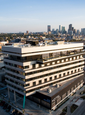 Aerial view of the Zeppelin at The Source Hotel, showcasing its modern architecture and surrounding green spaces. The image captures the vibrant rooftop area and beautifully landscaped grounds, emphasizing the hotel's urban environment and accessibility to nearby amenities. The blue sky and bright sunlight enhance the overall inviting atmosphere of the venue.