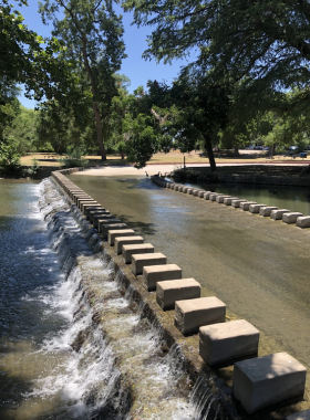 An old stone bridge with arched spans crosses a calm river, surrounded by trees. The bridge’s historic design blends harmoniously with the natural scenery, offering a tranquil, timeless view.