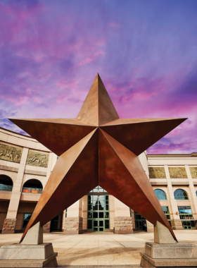 A stunning view of the Bullock Texas State History Museum, showcasing its modern architecture with large glass windows and a unique roof design. The entrance is adorned with Texas flags, inviting visitors to explore the state's rich history.

