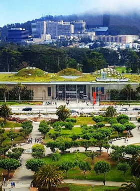 A striking exterior view of the California Academy of Sciences, showcasing its unique architecture with a green living roof adorned with native plants. The building features large glass panels that reflect the surrounding landscape, allowing natural light to flood the interior. Visitors can be seen entering the museum, highlighting its role as a popular destination for learning about science and nature in Golden Gate Park, San Francisco.

