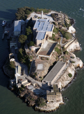 An aerial view of Alcatraz Island, featuring the historic prison building and surrounding waters. The rugged terrain and lush greenery contrast with the stark architecture of the prison, highlighting its historical significance.