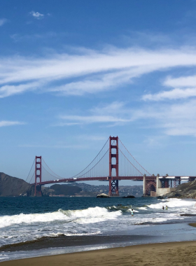 A stunning view of Baker Beach with the Golden Gate Bridge in the background. The sandy beach is dotted with sunbathers and beachgoers enjoying the warm sun, while gentle waves lap at the shore under a clear blue sky.