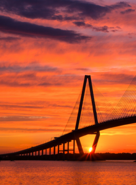 A serene sunset over the Charleston harbor, featuring a silhouetted fishing boat gently bobbing on the calm waters. The sky is ablaze with warm colors, including deep oranges, soft pinks, and golden yellows, creating a stunning backdrop for this tranquil scene. The reflection of the vibrant sky on the water enhances the beauty of the moment, embodying the peacefulness of a day ending in Charleston.

