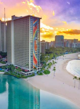 This image shows the stunning sunset over Waikiki Beach, with golden hues reflecting on the calm water, palm trees swaying gently in the breeze, and visitors enjoying the peaceful atmosphere on the sandy shore.