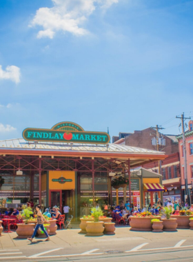 "This image shows a bustling scene at Findlay Market, with colorful fresh produce stalls offering a variety of local fruits, vegetables, and goods, drawing crowds of eager shoppers."

