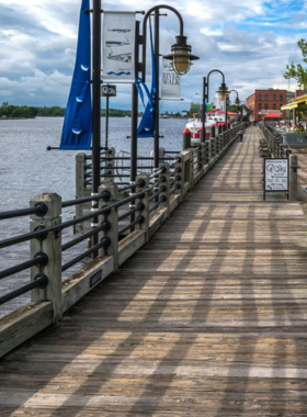 This image shows the picturesque Riverwalk in Wilmington, NC, offering a scenic view of the Cape Fear River with people strolling along the boardwalk, enjoying the beautiful waterfront.