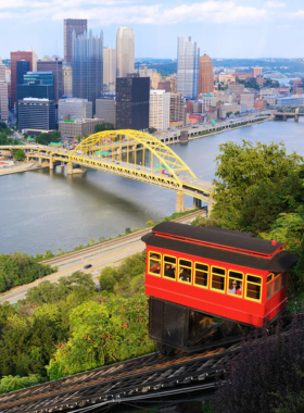 "This image shows the historic Duquesne Incline, a red-and-white cable car scaling the green hillside with Pittsburgh’s downtown skyline in the background. Visitors can see both the scenic incline and the city’s iconic view from Mount Washington."
