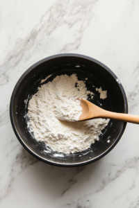 Flour sprinkled over softened veggies in black pot on white marble cooktop, stirred with a wooden spoon to create a smooth roux