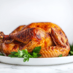 A beautifully roasted whole turkey is placed on a white ceramic serving tray atop a white marble countertop. The golden crispy skin, lightly brushed with olive oil and flavored butter, showcases a tender and juicy finish. The turkey is cooked to perfection and garnished with herbs, ready to be carved and served.