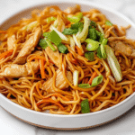 side view of the final chicken chow mein dish served on a white ceramic plate placed on a clean white marble countertop. The noodles are cooked perfectly and coated in a glossy sauce, surrounded by vibrant vegetables like cabbage, carrots, and green onions. Thin chicken strips are spread evenly, making the dish colorful and delicious. The composition is simple, highlighting the textures and colors of the completed chow mein.