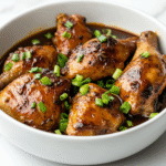 side view of the finished Filipino chicken adobo served in a clean white ceramic bowl. The chicken pieces are tender and glazed in a rich, dark brown sauce, generously garnished with freshly sliced green onions. The bowl is placed on a white marble countertop with a minimalistic setup, perfect for showcasing the vibrant, caramelized texture of the dish and making it appealing for any food enthusiast or home cook.