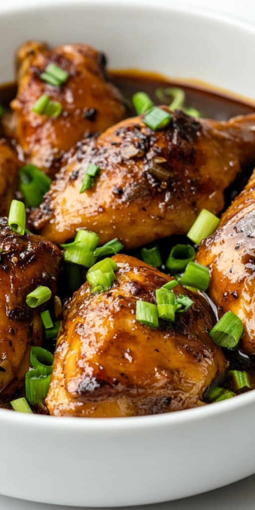 side view of the finished Filipino chicken adobo served in a clean white ceramic bowl. The chicken pieces are tender and glazed in a rich, dark brown sauce, generously garnished with freshly sliced green onions. The bowl is placed on a white marble countertop with a minimalistic setup, perfect for showcasing the vibrant, caramelized texture of the dish and making it appealing for any food enthusiast or home cook.