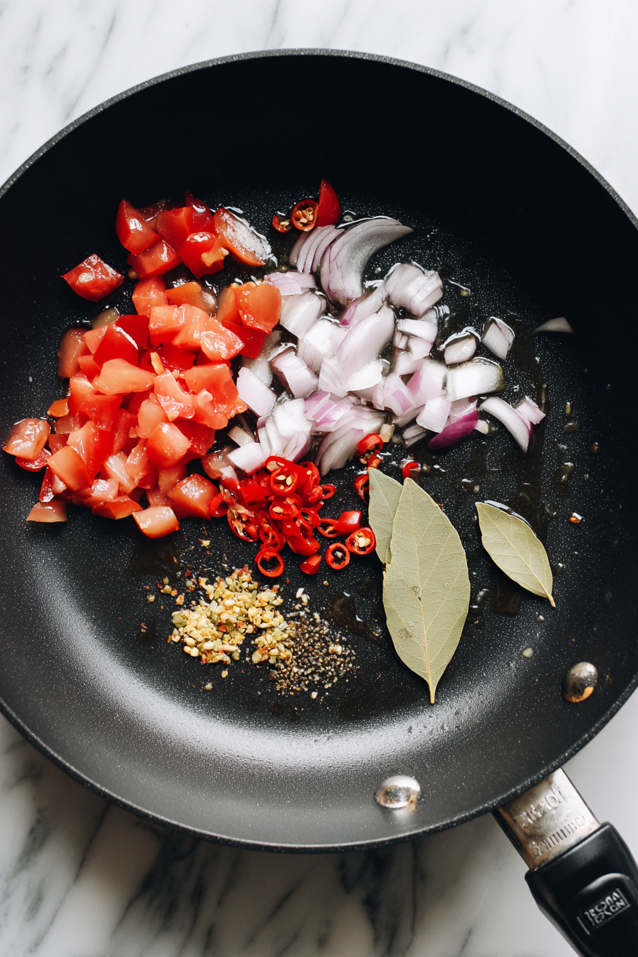 In a skillet, heat oil and sauté diced onion, tomato, garlic, red chili, and bay leaves until fragrant.